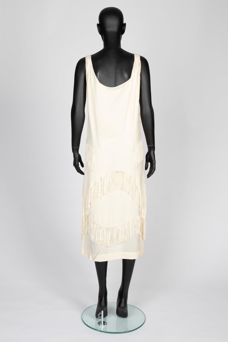 Ensemble i elfenbensfarget silkekrepp (crêpe georgette) med eggeskallhvite snorfrynser. Ensemblen består av tre deler; kjole, cape og skjerf. Kjolen er ermeløs, har rett fasong og vid utringning. Skjørtet er festet til et underliv, med en løsthengende bluse over, bare festet til underlivet bak. Blusen er tunget og har frynser nederst. Skjørtet har to løsthengende overskjørt eller kapper i forskjellig lengde, begge festet i livsømmen og med tunget avslutning og frynser. Capen er vid og i samme stoff, fôret med et bredt, kragelignende skjerf, som faller frem over skuldrene i lange, brede skjerfender med lange frynser. I ryggen to lange frynserader. Den nedre faller helt til capens nederkant. Langt skjerf med rundede avslutninger og frynser.