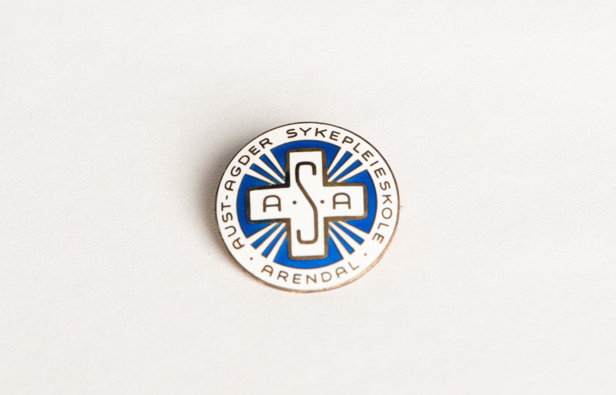 Pin/pyntenål brukt av sykepleier ved Aust-Agder Sykepleierskole, Arendal. Dekorert med hvit og blå emalje og lukkes med metallnål bak.