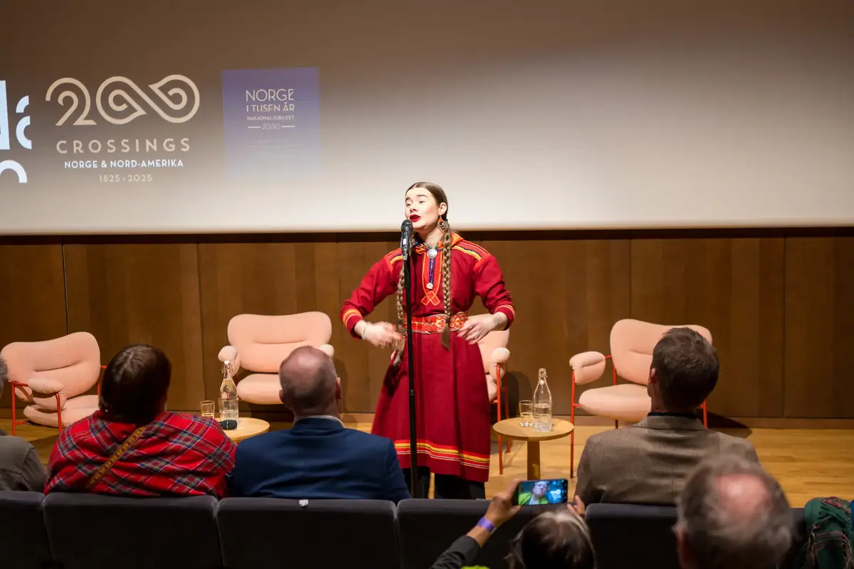 En kvinne med fletter i rød samekofte synger foran et publikum.