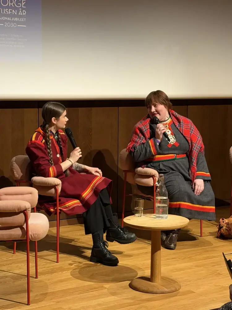 To kvinner i samekofter sitter i hver sin stol ved et lite bord snakker med hverandre med mikrofon i hendene.
