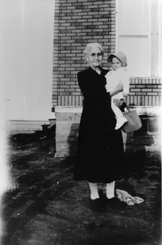 En eldre kvinne med briller mørk kjole holder et lite barn med lyse klær og en kyse på armen. På armen hennes henger også en håndveske. Ved hennes føtter ligger noe tøy. Hun står foran en murbygning.