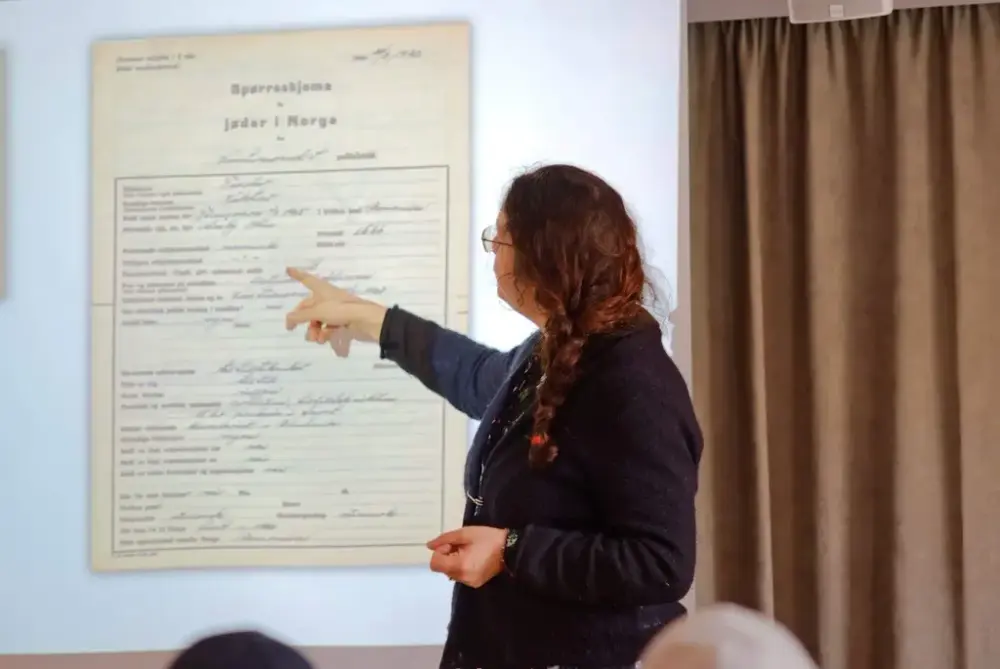 Bildet viser en kvinne med mørkt langt hår i flette, som står foran et lerret. Hun peker mot dokumentet på lerretet. 