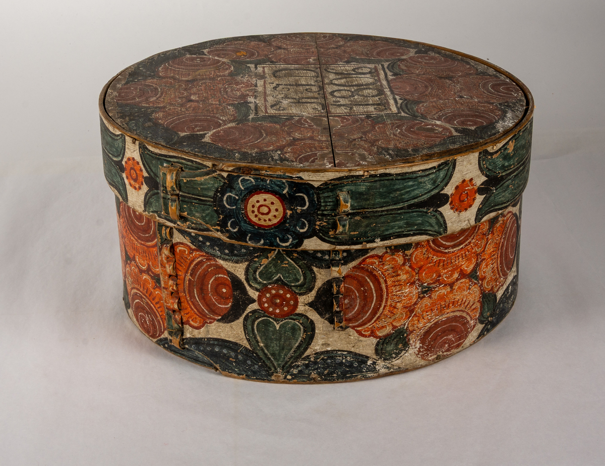 Cylindrisk, svept ask av björk, botten och lock av furu. Målat rosmotiv i rött och blått. Initialerna KJD samt årtalet 1806 på locket.