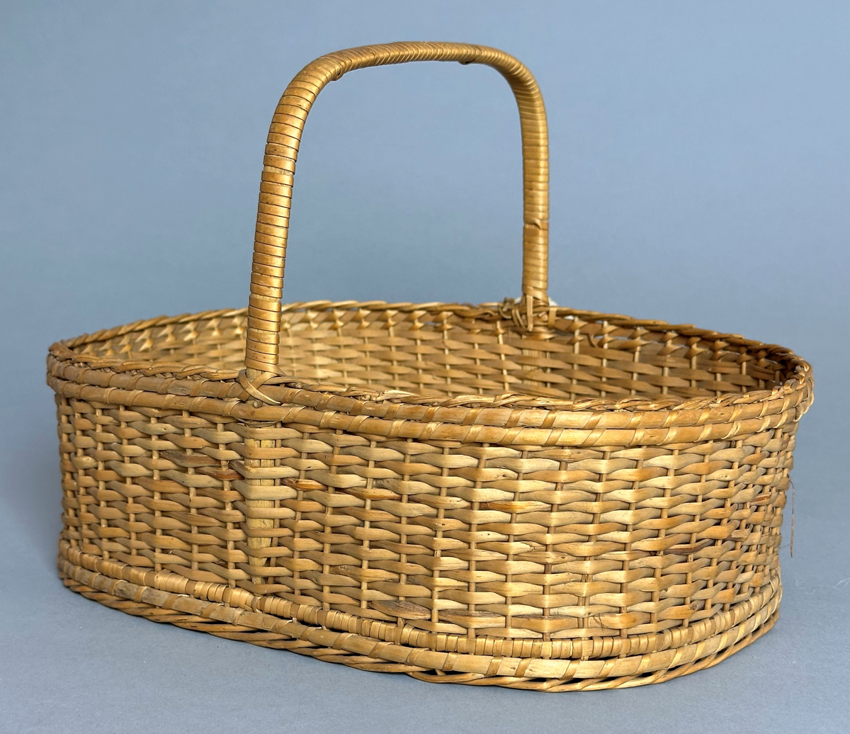 Rektangulär korg med rundade hörn, flätad i tuskaft av pil på böjda vidjor från botten till stående varp. Har ett lindat handtag mitt över av delad pil.