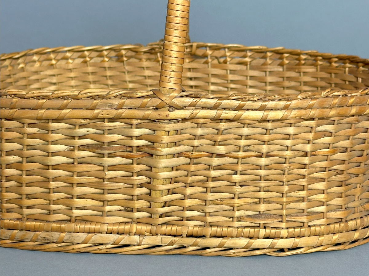 Rektangulär korg med rundade hörn, flätad i tuskaft av pil på böjda vidjor från botten till stående varp. Har ett lindat handtag mitt över av delad pil.