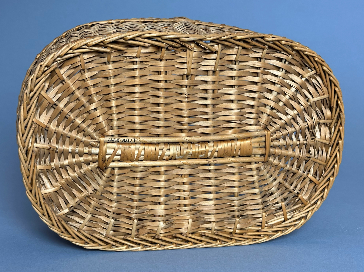 Rektangulär korg med rundade hörn, flätad i tuskaft av pil på böjda vidjor från botten till stående varp. Har ett lindat handtag mitt över av delad pil.
