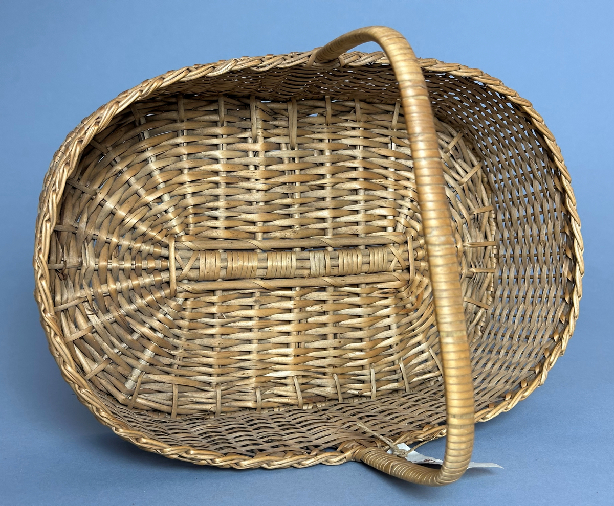 Rektangulär korg med rundade hörn, flätad i tuskaft av pil på böjda vidjor från botten till stående varp. Har ett lindat handtag mitt över av delad pil.