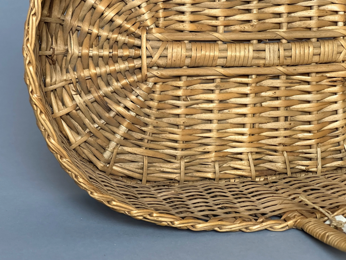 Rektangulär korg med rundade hörn, flätad i tuskaft av pil på böjda vidjor från botten till stående varp. Har ett lindat handtag mitt över av delad pil.