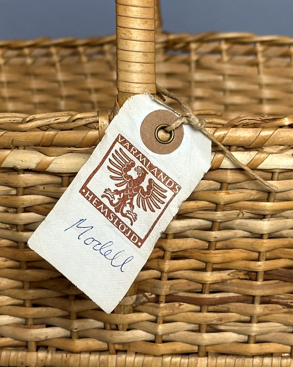 Rektangulär korg med rundade hörn, flätad i tuskaft av pil på böjda vidjor från botten till stående varp. Har ett lindat handtag mitt över av delad pil.