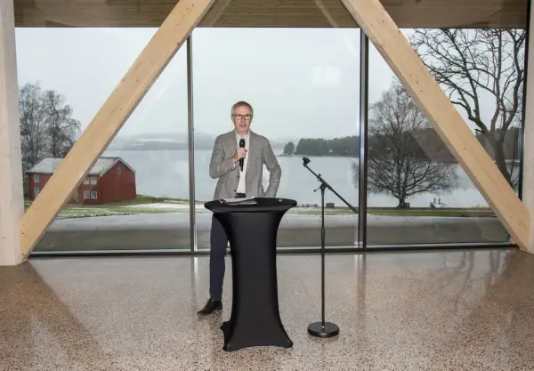 Øystein Nordal fra Martin M. Bakken står foran store vinduer, gjennom dem ses det røde huset fra Løtenprestegård og Mjøsa.