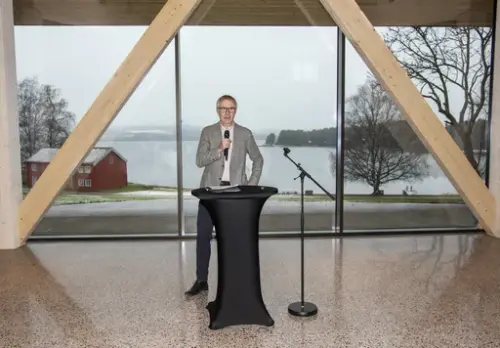 Øystein Nordal fra Martin M. Bakken står foran store vinduer, gjennom dem ses det røde huset fra Løtenprestegård og Mjøsa.