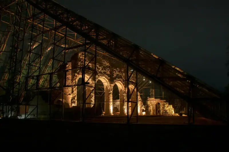 Middelalderruinene på Domkirkeodden er lyst opp i gulaktig lys, mens stålkonstruksjonen i vernebygget nærmest framstår som et fengsel i skarp kontrast. Mørk kveldshimmel rundt.