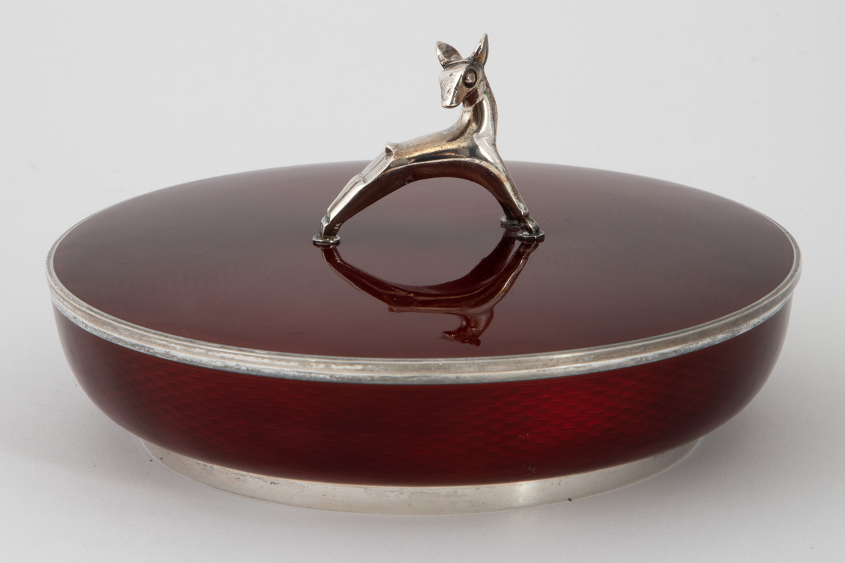 Rund rødemaljert sølvskål med lokk. Sølvbase og sølvkant på skålen og sølvkant på lokket. Grepet på lokket er formet som et dådyr. Dyp rød transparent emalje over et guillochert mønster av bølgende stipulerte linjer i sølvet.