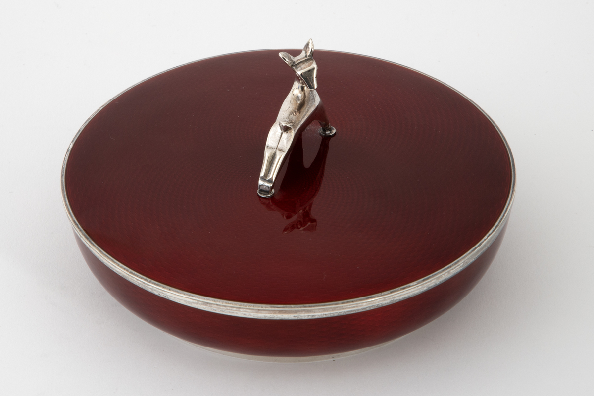 Rund rødemaljert sølvskål med lokk. Sølvbase og sølvkant på skålen og sølvkant på lokket. Grepet på lokket er formet som et dådyr. Dyp rød transparent emalje over et guillochert mønster av bølgende stipulerte linjer i sølvet.