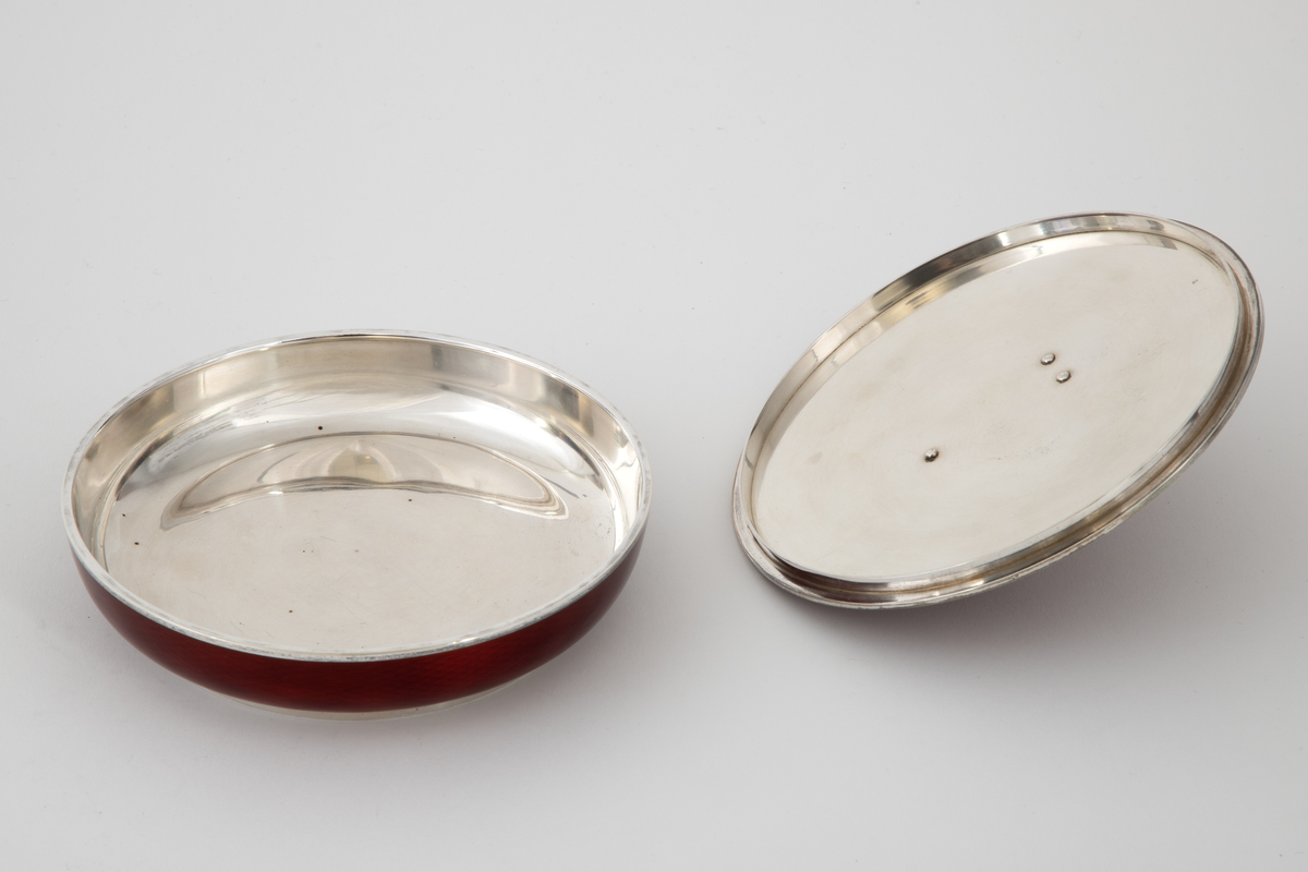 Rund rødemaljert sølvskål med lokk. Sølvbase og sølvkant på skålen og sølvkant på lokket. Grepet på lokket er formet som et dådyr. Dyp rød transparent emalje over et guillochert mønster av bølgende stipulerte linjer i sølvet.