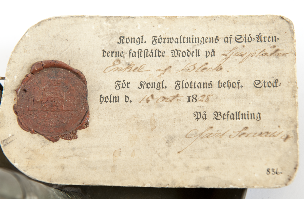 Modellexemplar av ljusstake, av bleckplåt med fat och hållare för vaxljus, 3 lägen, samt handtag. Vidhäftad etikett med sigill med texten "Kong. Förwaltningens af Sjö-Ärenderne faststälde modell på ljusplåtar enkel af bleck. För Kongl. Flottans behof. Stockholm d. 19 oct. 1828. På befallning Carl Servais"