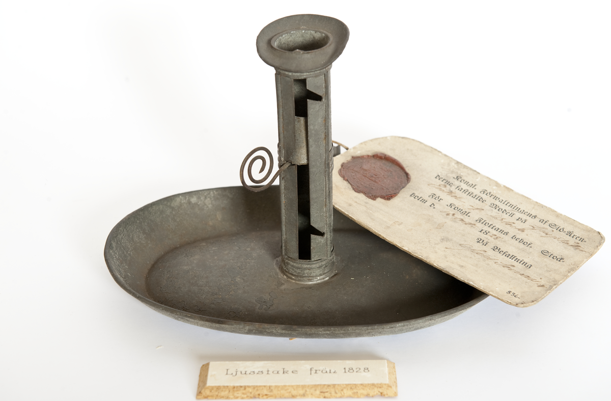 Modellexemplar av ljusstake, av bleckplåt med fat och hållare för vaxljus, 3 lägen, samt handtag. Vidhäftad etikett med sigill med texten "Kong. Förwaltningens af Sjö-Ärenderne faststälde modell på ljusplåtar enkel af bleck. För Kongl. Flottans behof. Stockholm d. 19 oct. 1828. På befallning Carl Servais"