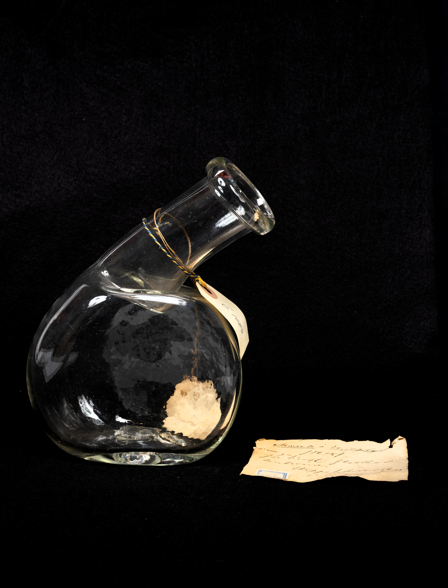 Modellexemplar av urinbägare i glas för flottans skeppsläkare. Med vidhäftad etikett. Lock saknas.