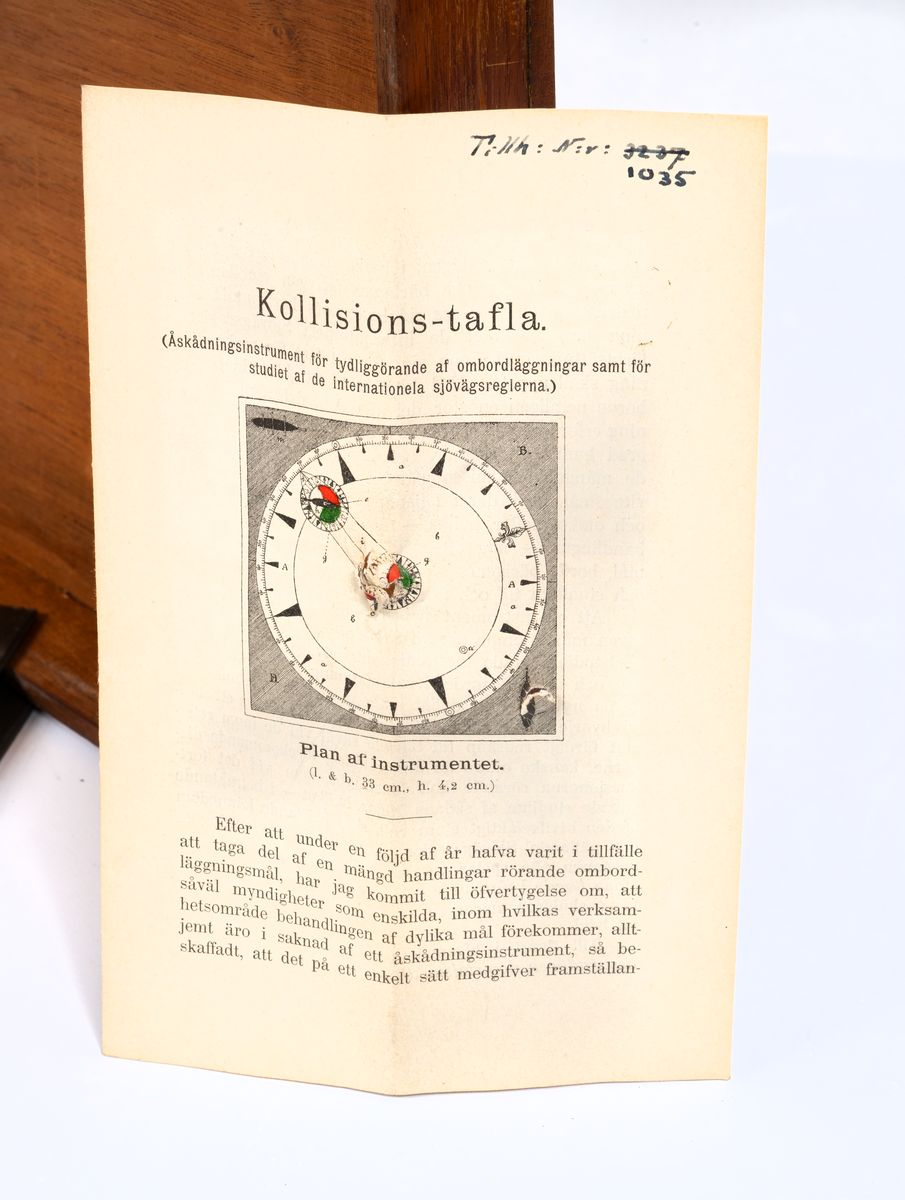 Kollisions-tavla, Ekelöfs patent, bestående av vit kompasssros med två rörliga farygsmodeller jämte visare, fästade till en rund bottenplatta, alltsammans monterat till en brunmålad, fyrkantig bottenplatta med lock av mahogny. Tillhörande tryckt beskrivning. Stockholm, 1892.