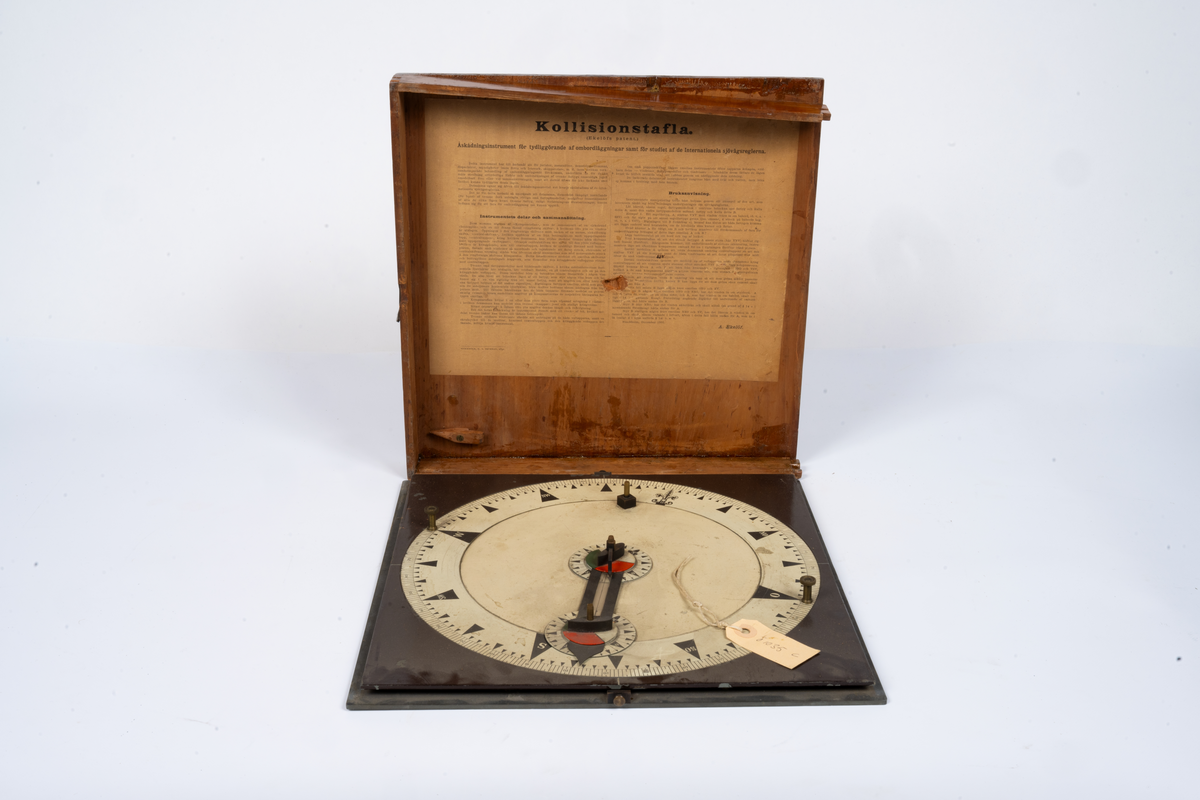 Kollisions-tavla, Ekelöfs patent, bestående av vit kompasssros med två rörliga farygsmodeller jämte visare, fästade till en rund bottenplatta, alltsammans monterat till en brunmålad, fyrkantig bottenplatta med lock av mahogny. Tillhörande tryckt beskrivning. Stockholm, 1892.