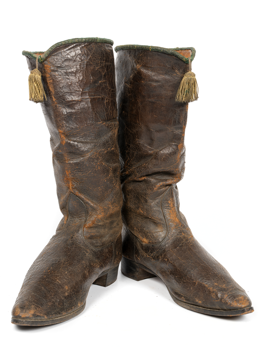 Stövlar, svartlackerade, korta skaft med guldtofs på framsidan, kantade med grönt läder. Sydda, smal fot och spetsig tå. Tillhörande järnskena för fästande till mannekäng.