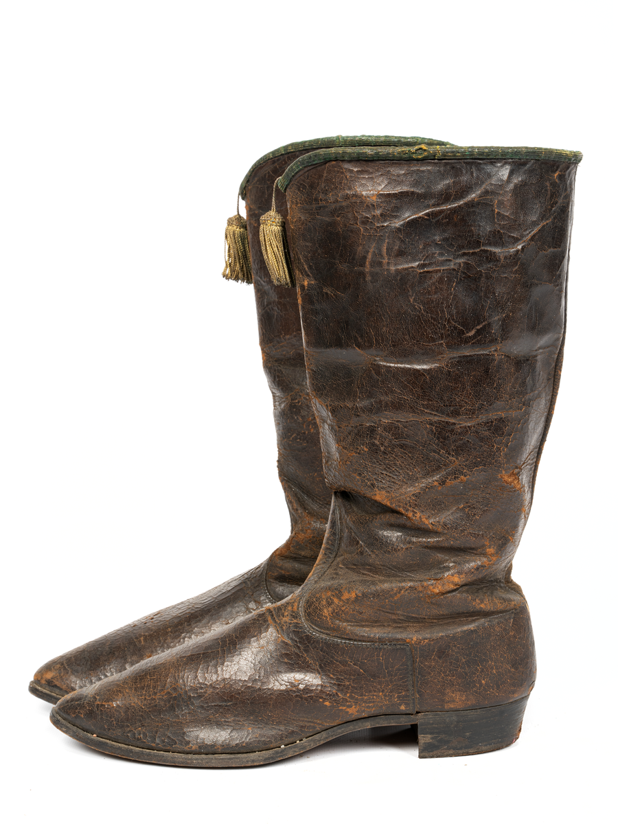 Stövlar, svartlackerade, korta skaft med guldtofs på framsidan, kantade med grönt läder. Sydda, smal fot och spetsig tå. Tillhörande järnskena för fästande till mannekäng.