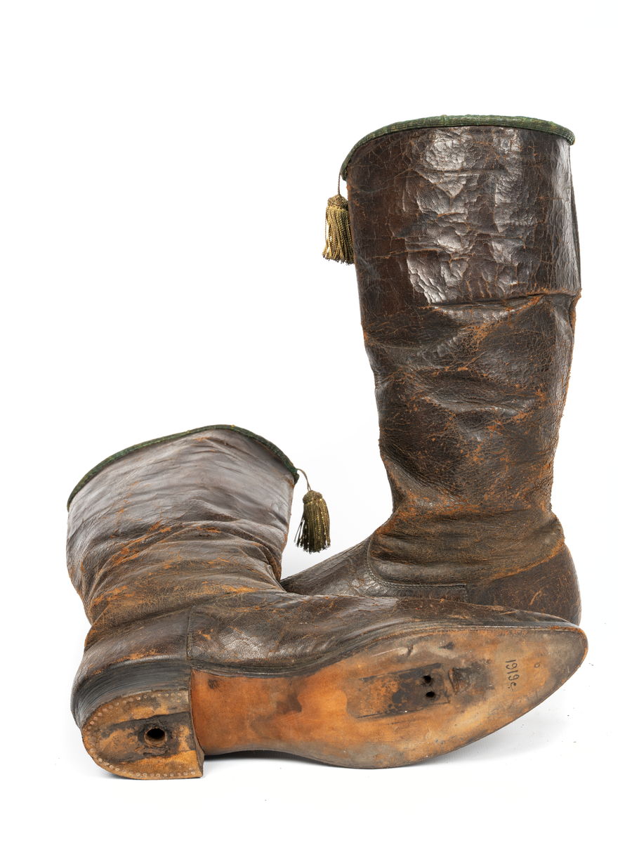 Stövlar, svartlackerade, korta skaft med guldtofs på framsidan, kantade med grönt läder. Sydda, smal fot och spetsig tå. Tillhörande järnskena för fästande till mannekäng.