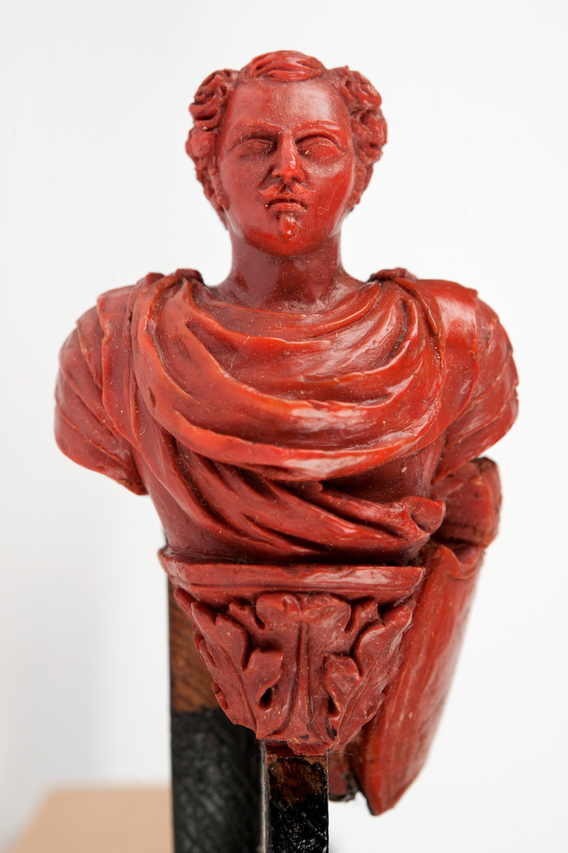 Förslag till galjon till PRINS OSCAR.
Bröstbild av Prins Oscar på sockel prydd med akantusblad och flankerad om babord med krönt sköld, skisserad. Monterad till stävkonstruktion av svartmålat trä. Röd vax, förslag