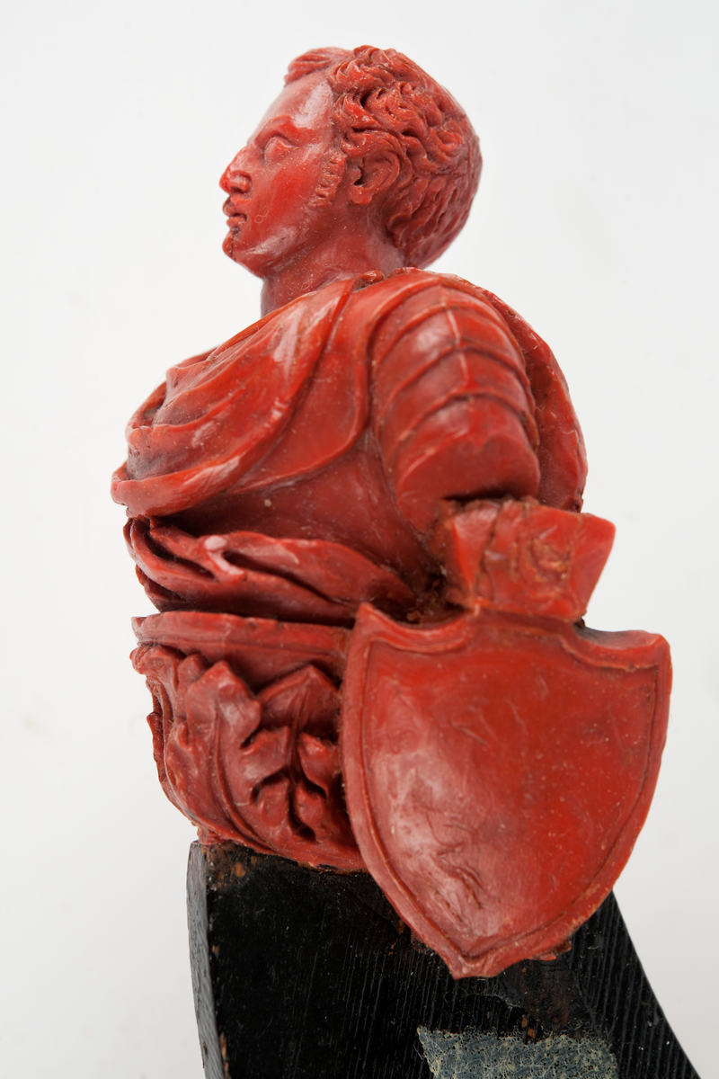 Förslag till galjon till PRINS OSCAR.
Bröstbild av Prins Oscar på sockel prydd med akantusblad och flankerad om babord med krönt sköld, skisserad. Monterad till stävkonstruktion av svartmålat trä. Röd vax, förslag