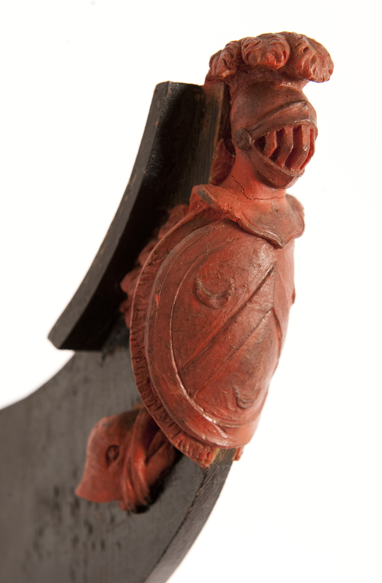 Föreställande fregatten AF CHAPMANs galjon. Vapensköld, oval med vinkel och tre halvmånar. Krönt av hjälm med fjäderbuske. På sidorna bladslinga samt om styrbord ett delfinhuvud. Monterat till stävkonstruktion av svartmålad träplatta, märkt ”Chapman”. 
Plattan: 155 x 85 mm röd vax