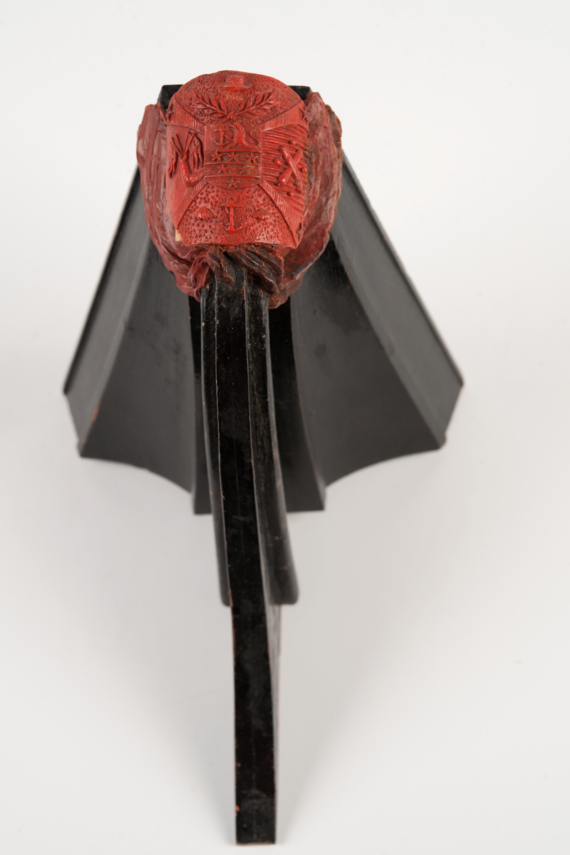 Föreställande korvetten LAGERBJELKEs vapensköld, omgiven av 2 flaggor på var sida. Monterad till stävkonstruktion av svartmålat trä med del av gråmålad bogspröt.
Röd vax.