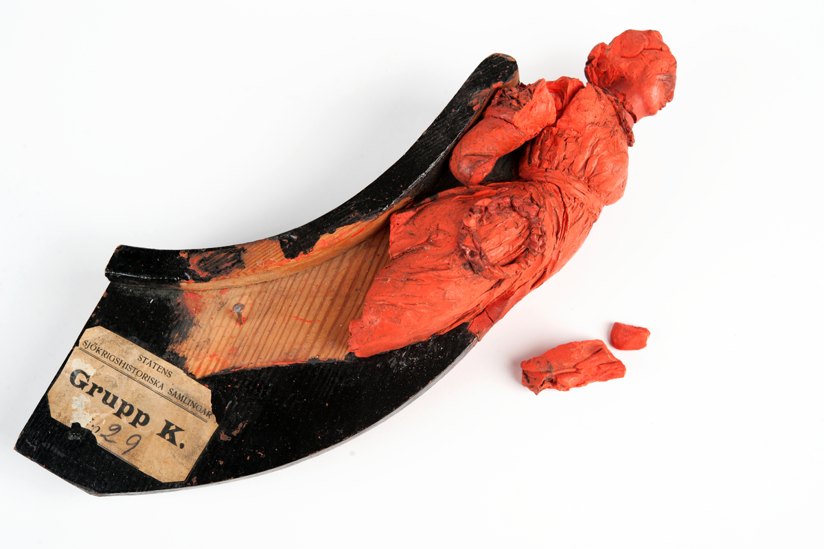 Fregatten DESIREEs galjon. Kvinnofigur i grekisk dräkt med en lagerkrans på högra sidan. Underarmar och fötter förlorade. Monterad till stävkonstruktion av svartmålat trä. Röd vax.