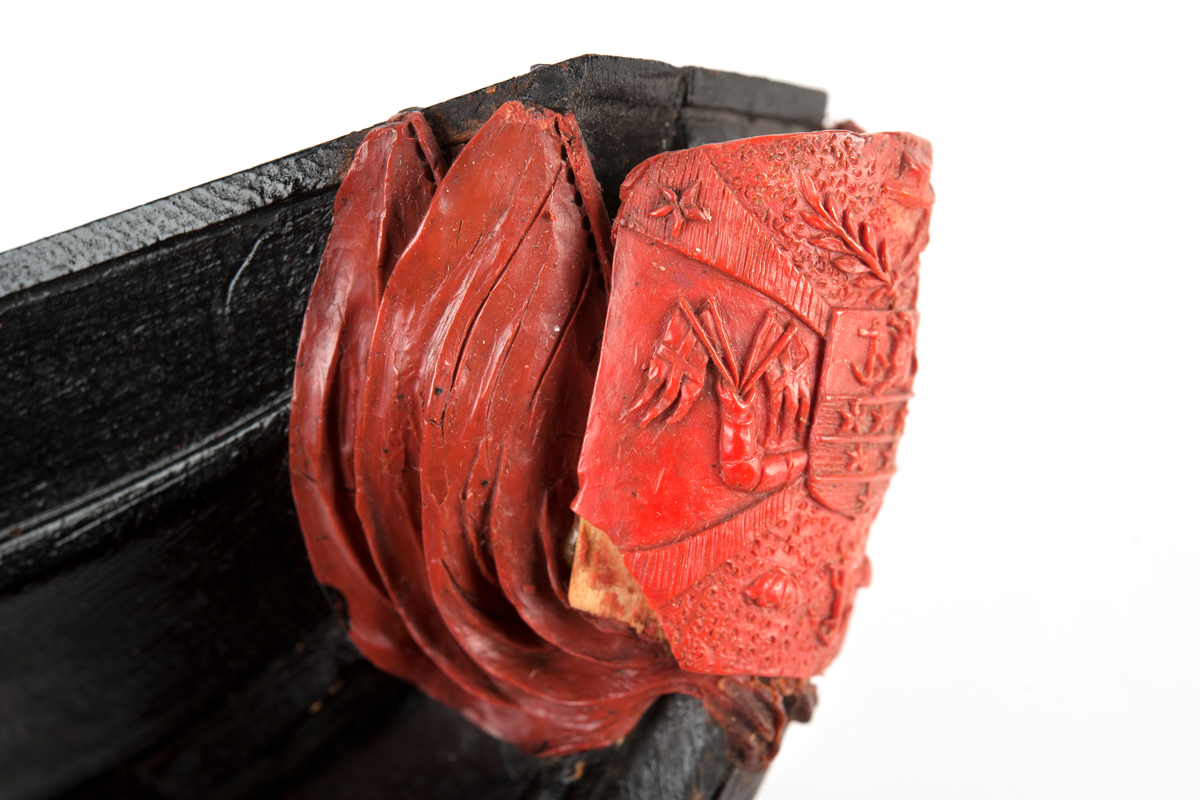 Föreställande korvetten LAGERBJELKEs vapensköld, omgiven av 2 flaggor på var sida. Monterad till stävkonstruktion av svartmålat trä med del av gråmålad bogspröt.
Röd vax.