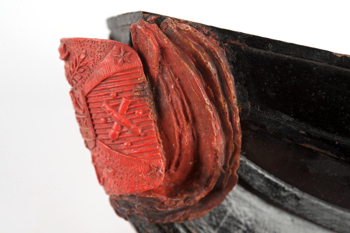 Föreställande korvetten LAGERBJELKEs vapensköld, omgiven av 2 flaggor på var sida. Monterad till stävkonstruktion av svartmålat trä med del av gråmålad bogspröt.
Röd vax.