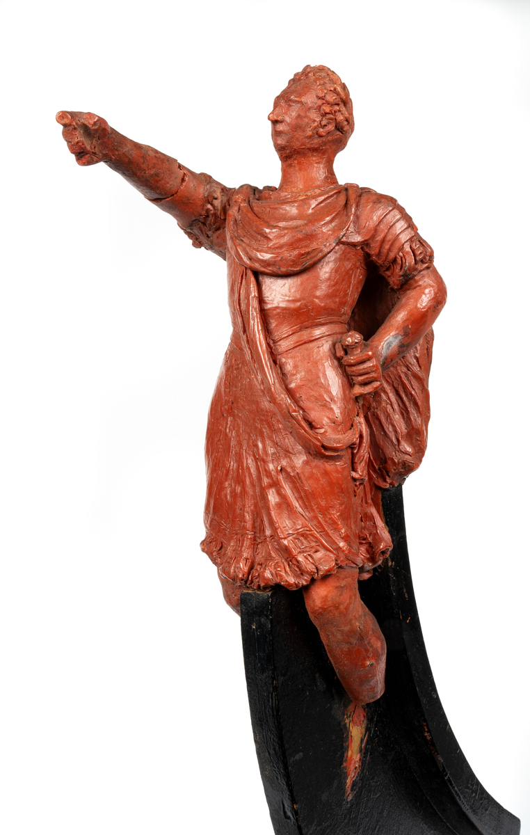 Förestäler Linjeskeppet Carl XIV Johans galjon. Mansfigur, Carl XIV Johan i  romersk dräkt. Höger arm avslagen. Monterad till
stävkonstruktion av svartmålat trä. Röd vax.