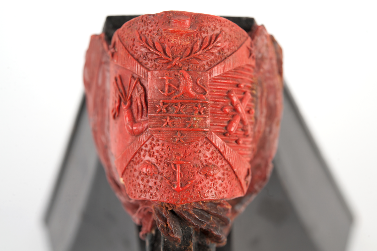 Föreställande korvetten LAGERBJELKEs vapensköld, omgiven av 2 flaggor på var sida. Monterad till stävkonstruktion av svartmålat trä med del av gråmålad bogspröt.
Röd vax.