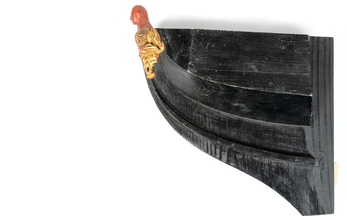 Korvetten EUGENIEs galjon. Kvinnobyst vilande på akantusblad. Nedtill om styrbord bladslinga. Huvudet skadat. Dräkt och blad förgyllda men röda vaxet synas. Monterad till stävkonstruktion av svartmålat trä. Röd vax.