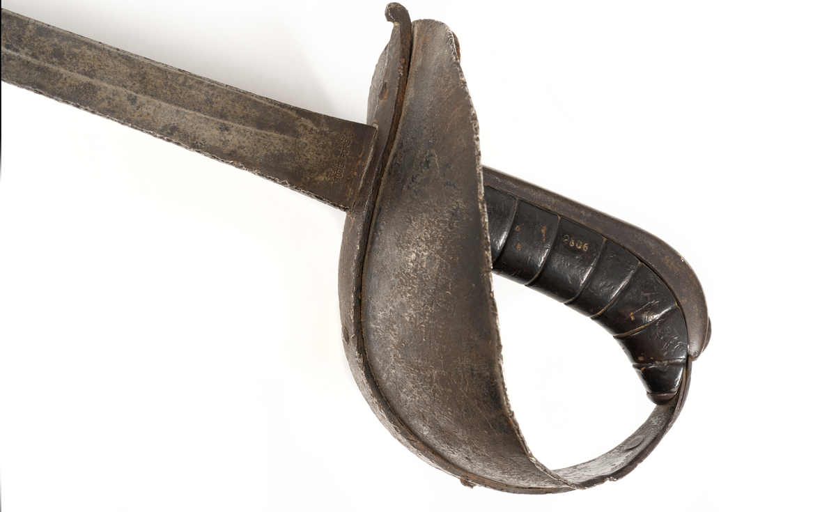 Fältsabel M/1860. Rak, jämnsmal klinga med rund spets. Eneggad med en bred blodränna närmast ryggen. Eggen är naggad. Fäste med stor  symetrisk parerplåt av järn. Kavel läderklädd med rygg av järn, samt handskydd av läder.
