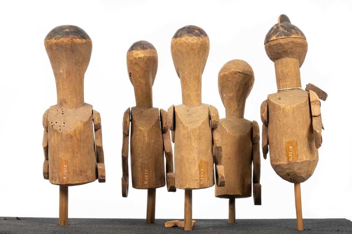 Modell av kanot från Samoa, skuren ur en stam av balsaliknande trä med relingen urhakad för tio tvärsgående balkar. Till modellen hör 5 snidade mansfigurer, fem paddlar, en fågel och fyra olika snidade tofter eller dylikt. Flera smärre skador. Större skada i aktern.