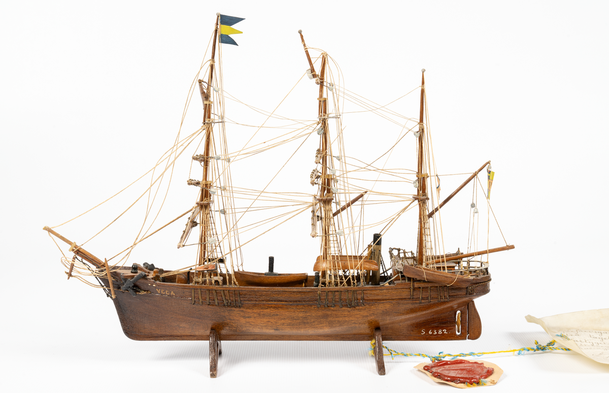 Fartygsmodell, hel i block av ek, taget från fartygets köl, expeditionsfartyget VEGA, barkriggat, utan segel, tvåbladig propeller, hög vit skorsten med svart "top". Skrå av ek, propeller och drill av ben. KSSS standert, tretungad flagga med 0 II:s namnchiffer. Äkthetsintyg ang. virket på pergament med vidfäst sigill daterat ll/3 1884, "Warfvet Kusten", Göteborg. Glaslåda.