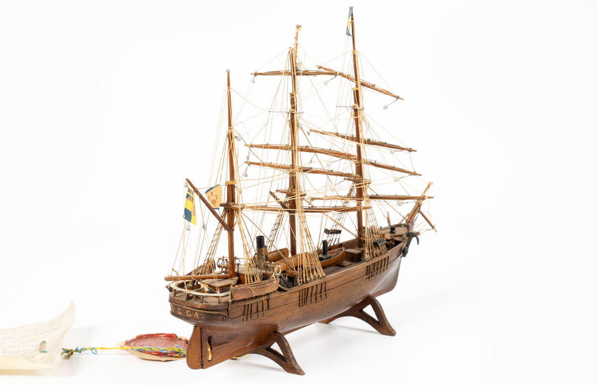 Fartygsmodell, hel i block av ek, taget från fartygets köl, expeditionsfartyget VEGA, barkriggat, utan segel, tvåbladig propeller, hög vit skorsten med svart "top". Skrå av ek, propeller och drill av ben. KSSS standert, tretungad flagga med 0 II:s namnchiffer. Äkthetsintyg ang. virket på pergament med vidfäst sigill daterat ll/3 1884, "Warfvet Kusten", Göteborg. Glaslåda.