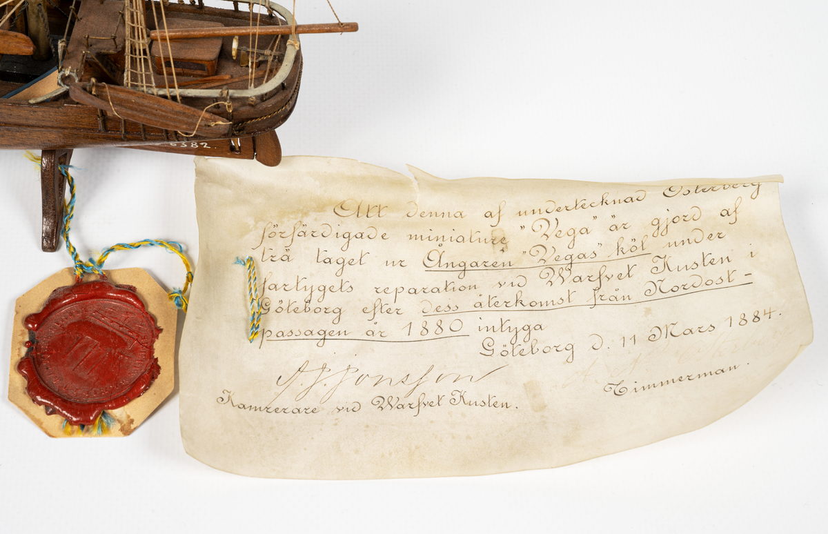 Fartygsmodell, hel i block av ek, taget från fartygets köl, expeditionsfartyget VEGA, barkriggat, utan segel, tvåbladig propeller, hög vit skorsten med svart "top". Skrå av ek, propeller och drill av ben. KSSS standert, tretungad flagga med 0 II:s namnchiffer. Äkthetsintyg ang. virket på pergament med vidfäst sigill daterat ll/3 1884, "Warfvet Kusten", Göteborg. Glaslåda.