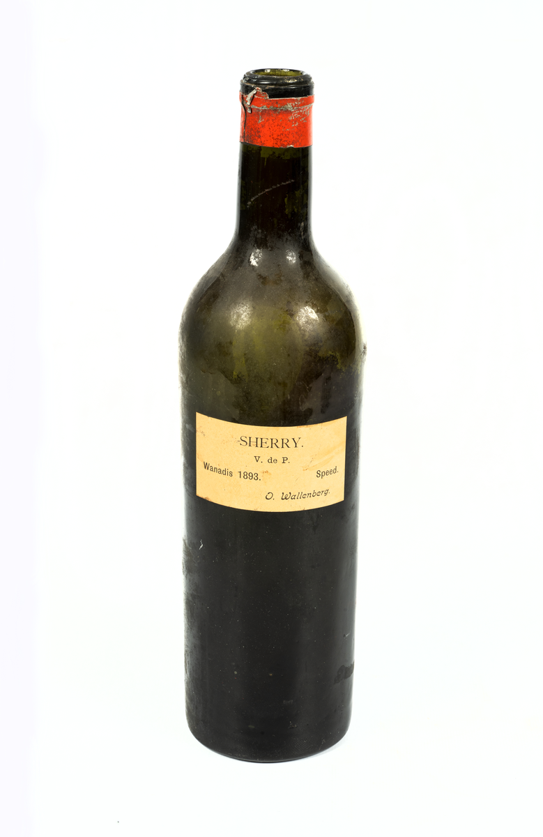 Flaska, tomglas, utan kork med kvarsittande röd kapsyl kring halsen.