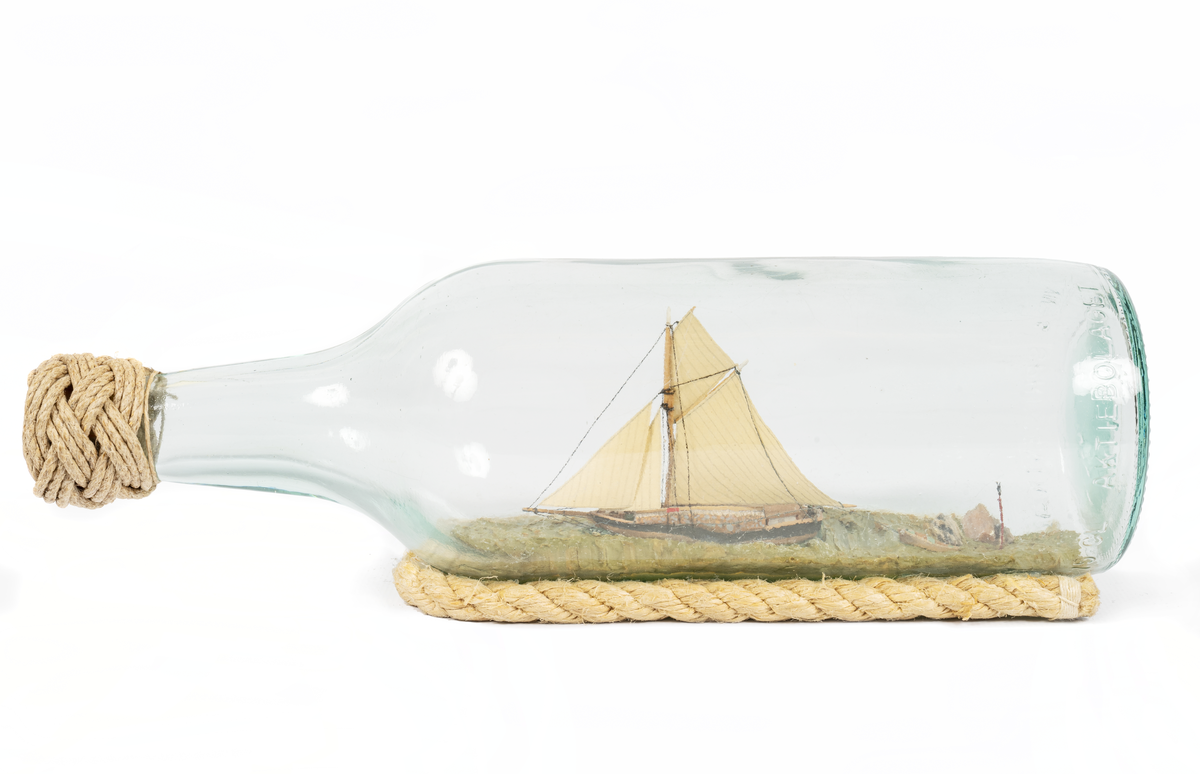 Flaskskepp av roslagsskuta under segel med gaffelstorsegel, gaffeltoppsegel, fock, klyvare. Jolle på släp. Skrå av otjärat rep.