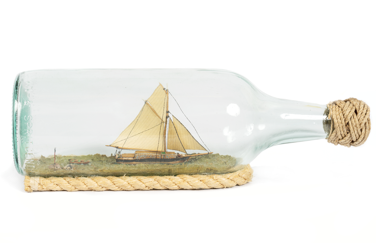 Flaskskepp av roslagsskuta under segel med gaffelstorsegel, gaffeltoppsegel, fock, klyvare. Jolle på släp. Skrå av otjärat rep.