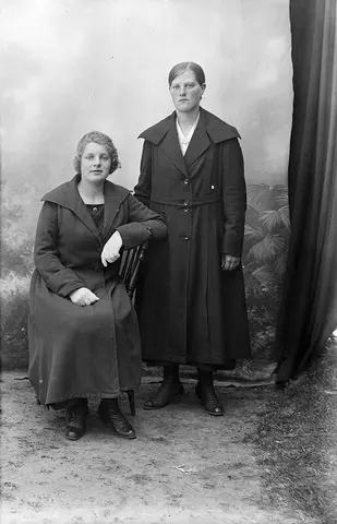 Portrett av to kvinner, den ene sitter på en stol og den andre står ved siden av. Kvinnene har kåpe på seg.