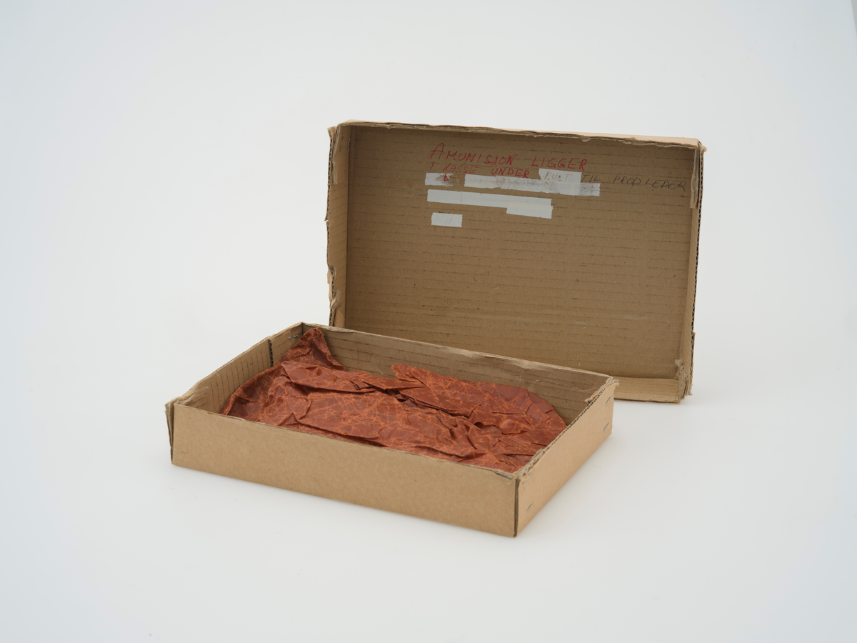 Eske med lokk til oppbevaring av signalpistol. Esken er en del av den originale emballasjen og har informasjon om pistolen stemplet på. Tekst og beskjeder om bruk er skrevet på for hånd flere steder på esken. Innvendig i esken ligger innpakningspapir som sannsynligvis er en del av den originale emballasjen.