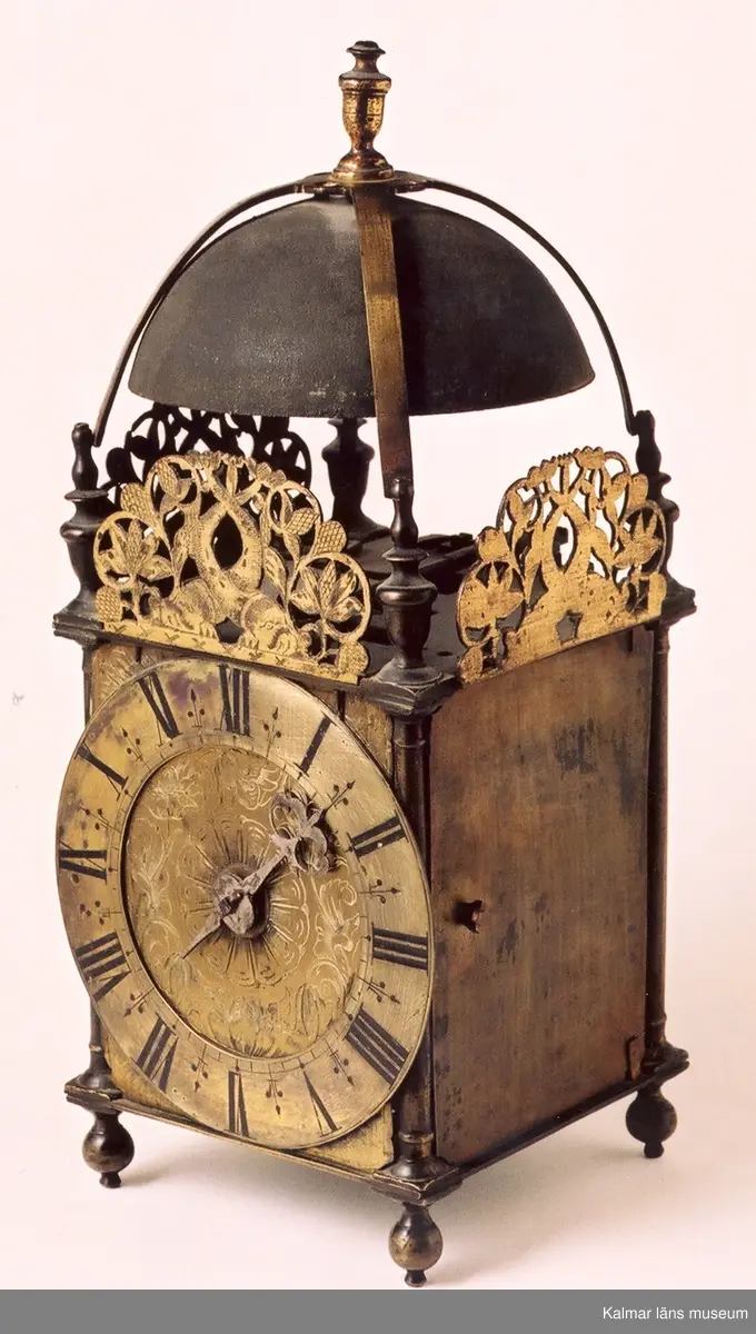 KLM 39255:145. Bordsur, lyktur, konsolur. Av mässing och järn. Krona och korsade metallband. Genombrutna brännförgyllda mässingsplattor på tre av sidorna. Den främre med graverat delfin- och blomstermotiv. Rund brännförgylld, graverad urtavla med tulpandekor. Sifferring fäst utanpå tavlan. Fyra kulfötter. Urverket är av mässing. Urverket är ej komplett, bl.a. saknas lod, lodlinor och pendel. Tillhörande konsolhylla i brunbetsad ek med urtag för lod och pendel. Profilerade sidostycken samt bakstycke dekorerat med rom-och fyrpassformade urtag. Upphängningsbeslag i mässing.