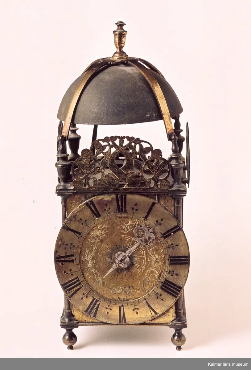 KLM 39255:145. Bordsur, lyktur, konsolur. Av mässing och järn. Krona och korsade metallband. Genombrutna brännförgyllda mässingsplattor på tre av sidorna. Den främre med graverat delfin- och blomstermotiv. Rund brännförgylld, graverad urtavla med tulpandekor. Sifferring fäst utanpå tavlan. Fyra kulfötter. Urverket är av mässing. Urverket är ej komplett, bl.a. saknas lod, lodlinor och pendel. Tillhörande konsolhylla i brunbetsad ek med urtag för lod och pendel. Profilerade sidostycken samt bakstycke dekorerat med rom-och fyrpassformade urtag. Upphängningsbeslag i mässing.