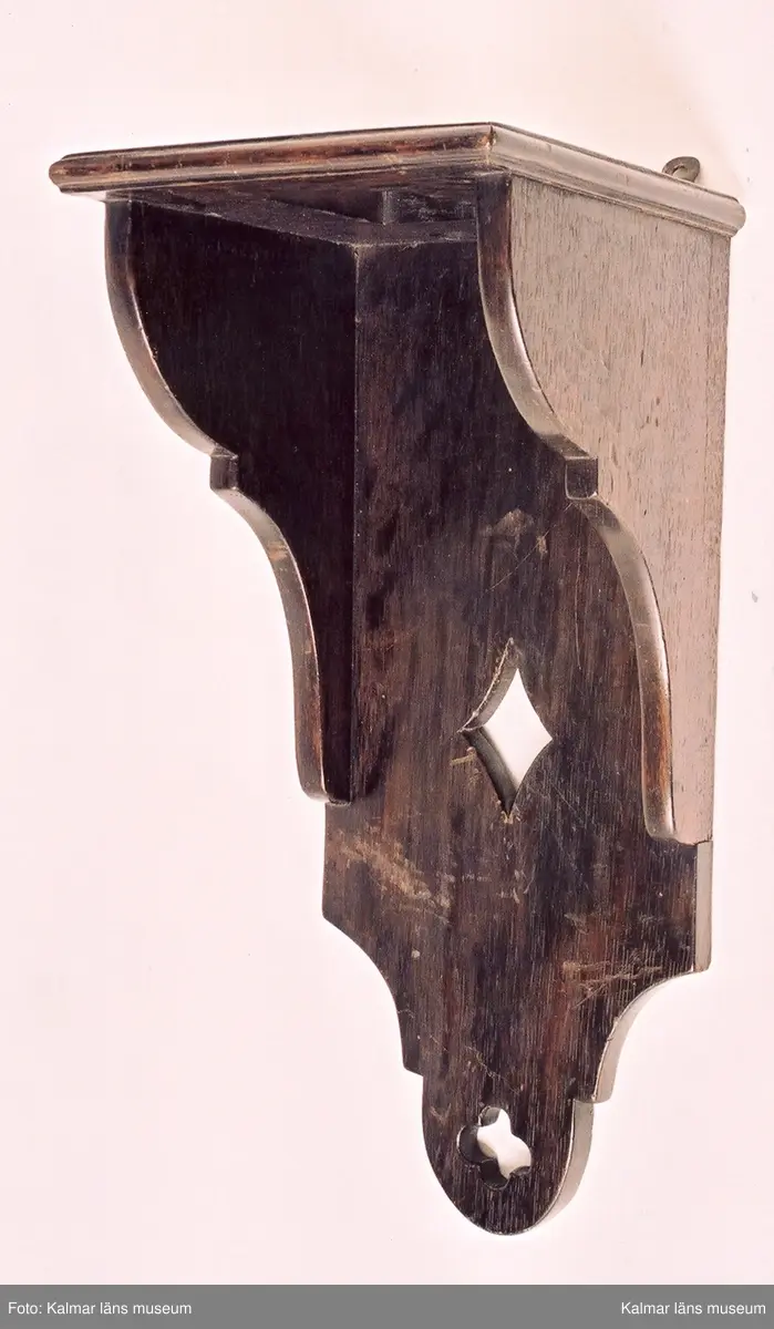 KLM 39255:145. Bordsur, lyktur, konsolur. Av mässing och järn. Krona och korsade metallband. Genombrutna brännförgyllda mässingsplattor på tre av sidorna. Den främre med graverat delfin- och blomstermotiv. Rund brännförgylld, graverad urtavla med tulpandekor. Sifferring fäst utanpå tavlan. Fyra kulfötter. Urverket är av mässing. Urverket är ej komplett, bl.a. saknas lod, lodlinor och pendel. Tillhörande konsolhylla i brunbetsad ek med urtag för lod och pendel. Profilerade sidostycken samt bakstycke dekorerat med rom-och fyrpassformade urtag. Upphängningsbeslag i mässing.